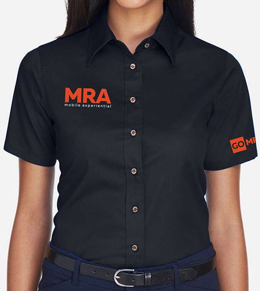 A smiling woman wearing a black short-sleeve button-up shirt with orange 'MRA Media Association' and 'GO MRA' logos, paired with a black belt, against a light gray background.